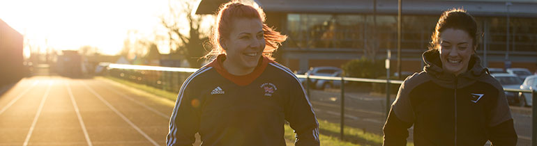 Students running track at Walsall Campus, both wearing sport jackets, the sun setting behind them